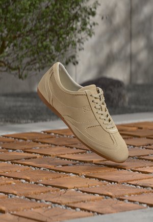 Baskets en daim beige à lacets flottant au-dessus d'un sol en carrelage en bois, avec de la verdure et un mur gris en arrière-plan.