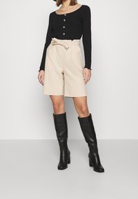 Svart långärmad topp med rund halsringning och knapppdetaljer, i kombination med beigefärgade högmidjade shorts och svarta högklackade stövlar som går upp till knäna.