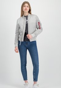 Ljusgrå bomberjacka med ribbad mudd och nederkant, som har en siddragkedja och en patch på vänster ärm, kombinerad med blå jeans och vita sneakers.