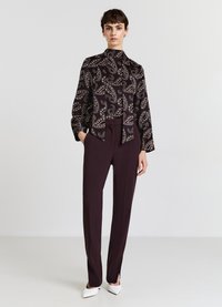 Blouse noire à imprimé paisley avec un col à nouer, associée à un pantalon bordeaux ajusté et des escarpins blancs à bout pointu. Tissu lisse, coupe structurée.