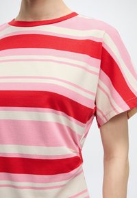 Άτομο που φοράει κοντομάνικο πουκάμισο με οριζόντιες ρίγες σε κόκκινο, ροζ και εκρού και μικρό κεντημένο λογότυπο "MODSTRØM" στο στήθος.