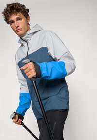 Veste légère tricolore en gris, marine et bleu. Dotée d'une capuche, d'une fermeture éclair à l'avant et de poignets réglables. Comprend des bâtons de trekking en main.