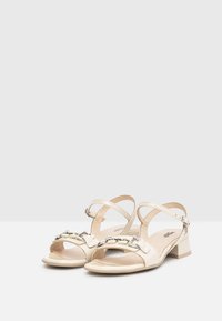 Sandali in pelle lucida beige con tacco a blocco, caratterizzati da una fascia ampia con accento in catena argentata e cinturini alla caviglia regolabili.
