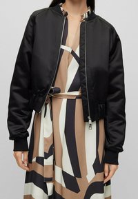 Svart kort bomberjacka i slät satin med ribbade mudd och krage, med dragkedja framtill och matta mönster.