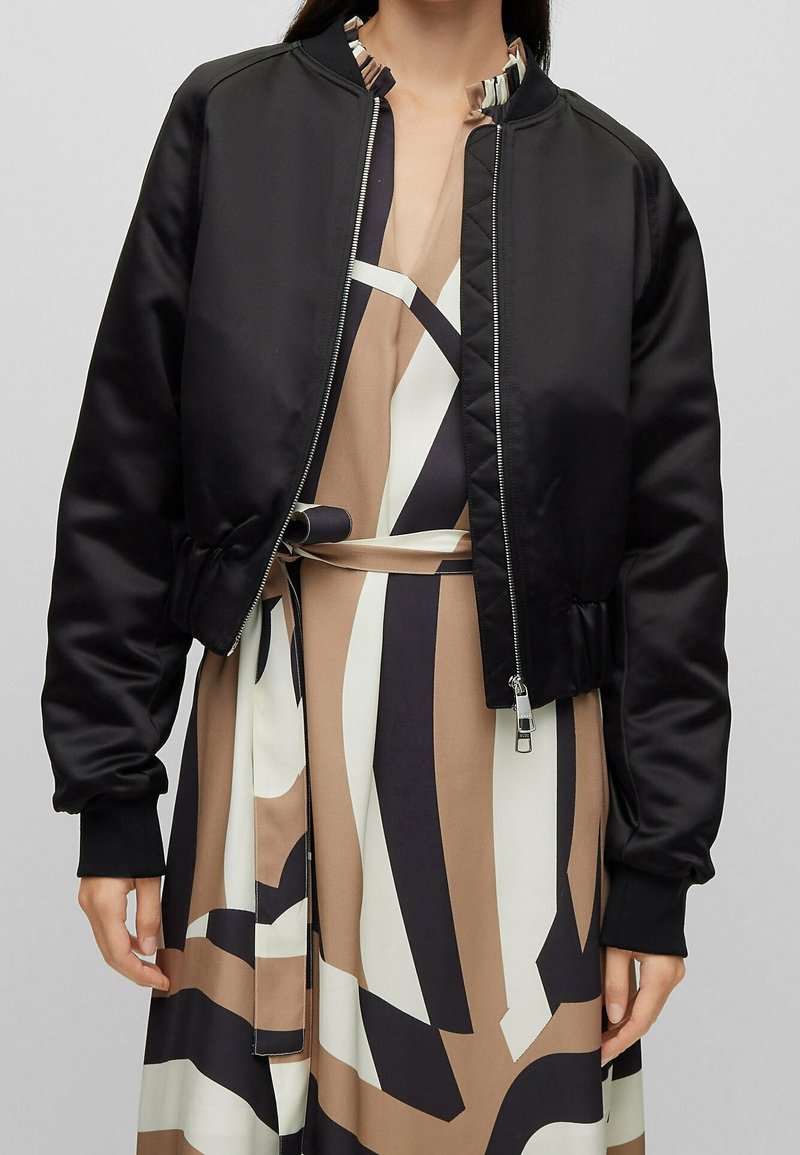 Svart kort bomberjacka i slät satin med ribbade mudd och krage, med dragkedja framtill och matta mönster.