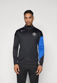 Black zip-up jacket with a high collar, featuring a blue left sleeve and a logo on the chest. Material appears smooth and athletic.