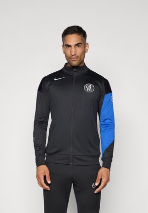 Black zip-up jacket with a high collar, featuring a blue left sleeve and a logo on the chest. Material appears smooth and athletic.