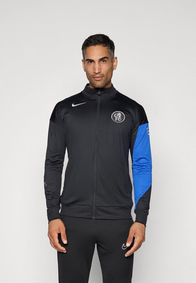 Black zip-up jacket with a high collar, featuring a blue left sleeve and a logo on the chest. Material appears smooth and athletic.
