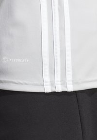 Lichtgrijs sporttop met drie witte verticale strepen, gemaakt van glad materiaal met het "AEROREADY" logo op de linkeronderzijde. Zwarte stof eronder.