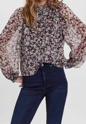 Femme portant un chemisier à manches longues à imprimé fleuri avec des manches bouffantes transparentes et un jean foncé taille haute sur un fond uni.