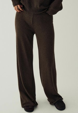 Personne portant un pantalon ample en maille marron foncé et un pull assorti avec des chaussures foncées sur un fond neutre.