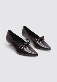 Zapatos de punta en piel sintética brillante de color marrón oscuro, con un pequeño tacón y lazos decorativos con acentos dorados en la parte frontal.