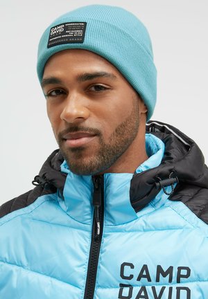 Light blue padded jacket with black accents, high collar, and zipper; paired with a light blue ribbed knit beanie featuring a brand patch.
