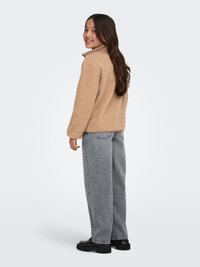 Ung person med langt hår iført en beige fleecejakke, løse grå jeans og sorte sko, stående og smilende over den ene skulder.