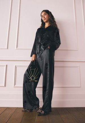 Femeie în costum pantaloni din satin negru cu modele florale, ținând o geantă unghiulară neagră cu design stea aurie, purtând pantofi cu toc ascuțit cu imprimeu leopard.