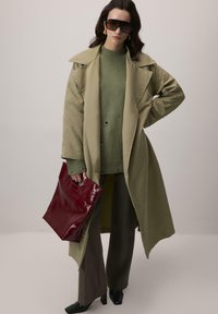 Zielona oliwkowa trenchcoat, dzianinowy sweter z dziurami, brązowe szerokie spodnie, błyszcząca bordowa torebka, czarne buty na obcasie, oversize'owe okulary przeciwsłoneczne.