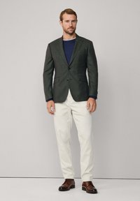 Tmavozelený blejzer s dvoma gombíkmi, nosený cez modré tričko s okrúhlym výstrihom, v kombinácii s bielymi nohavicami a hnedými šnurovacími topánkami. Jednoduchá štruktúra.