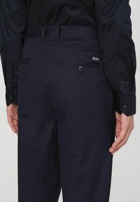 Pantaloni blu navy con una consistenza liscia, tasche posteriori con chiusure a bottone e un'etichetta di marca visibile cucita sul lato destro.