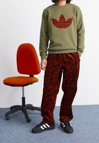 Olivgrön sweatshirt med stort röd mönsterlogotyp, i kombination med svarta och röda mönstrade byxor, bärs med svarta skor med vita ränder.