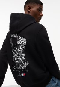 Ung mann iført svart hettegenser med hvit racerbile, sjakkbrettmønster og "Cadillac Formula 1 Team" grafikk på baksiden, kombinert med blå jeans.