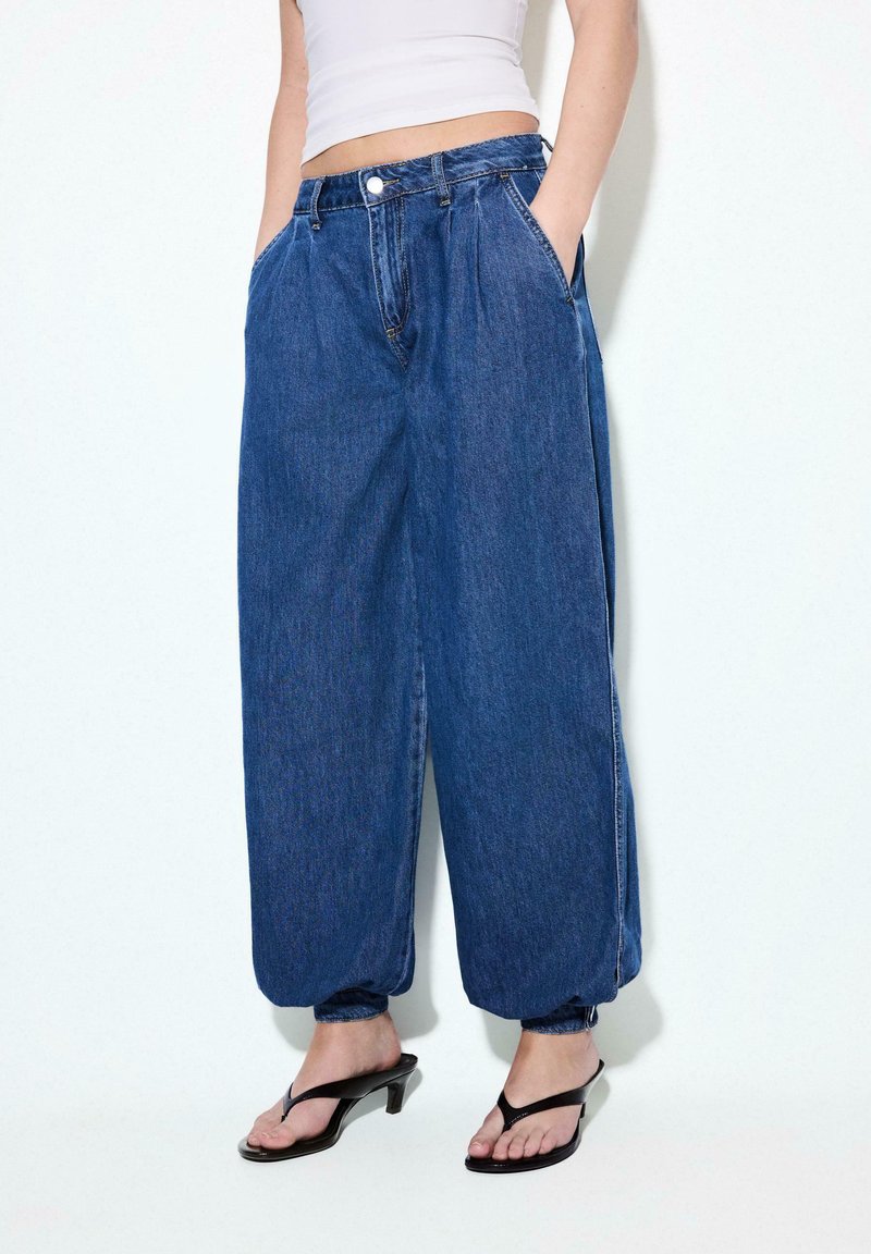 Personne portant un pantalon en denim bleu foncé à coupe ample avec des chevilles élastiquées, un haut court blanc, et des sandales à talons noires. Mains dans les poches.