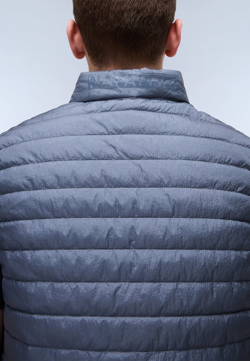 Szürke puffervest, mely puha tapintású, vízszintes steppelésű és álló gallérral rendelkezik. A logó a nyaktájékon látható.