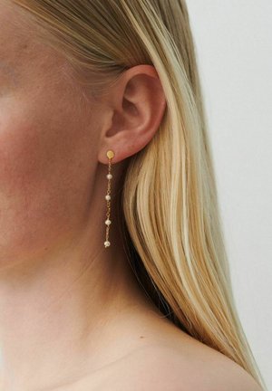 Personne blonde portant une longue boucle d'oreille en chaîne dorée avec cinq petites perles, montrant l'oreille gauche et le côté du cou sur un fond blanc.