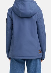 Zadní pohled na osobu oblečenou v modré kapucí a modrých džínech na jednobarevném pozadí.