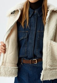 Crèmekleurige schapenvachten jack met een ritssluiting, met een zachte binnenkant, en een spijkerblouse eronder. Bruine riem met een gouden gesp op donkerblauwe denim.