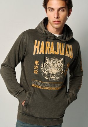 Joven con sudadera con capucha Harajuku verde oscuro con gráfico de tigre y caracteres japoneses, manos en los bolsillos delanteros, fondo neutro.