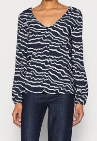 Donna che indossa una blusa a bottoni blu navy con un motivo a strisce ondulate bianche e maniche lunghe, abbinata a jeans blu scuro.