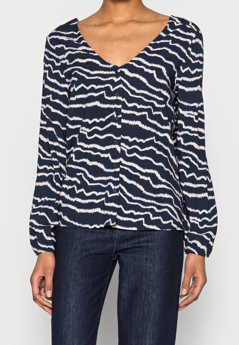 Donna che indossa una blusa a bottoni blu navy con un motivo a strisce ondulate bianche e maniche lunghe, abbinata a jeans blu scuro.