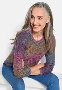 Femme souriante avec de longs cheveux gris, portant un pull en maille multicolore et un jean bleu, assise et regardant sur le côté.