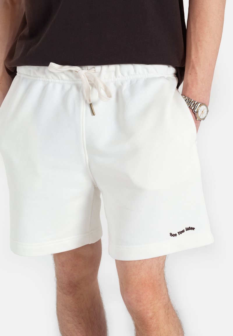 Białe bawełniane szorty z elastycznym pasem na sznurku, z małym haftowanym napisem "Do zobaczenia później" w ciemnym kolorze. Luźny fason, miękka tekstura.