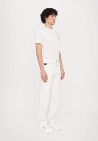 T-shirt en coton blanc avec un logo circulaire, associé à un jean en denim blanc et des baskets blanches, mettant en avant une coupe ajustée et un design minimaliste.