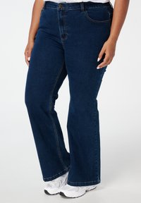 Donkerblauwe denim jeans met een rechte pijp, voorzien van vijf zakken, riemlussen en contrasterende stiksels. Draagt met witte sneakers.