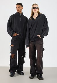 Tmavošedý oversized pulóver s golierom na zips, skombinovaný s roztrhnutými čiernymi nohavicami s veľkými dierami a béžovými záplatami, doplnený čiernymi čižmami.
