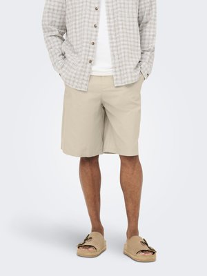 Homme portant un short beige mi-cuisses, des sandales ouvertes beiges avec des boucles, une chemise blanche et une chemise claire à carreaux boutonnée, les mains dans les poches.