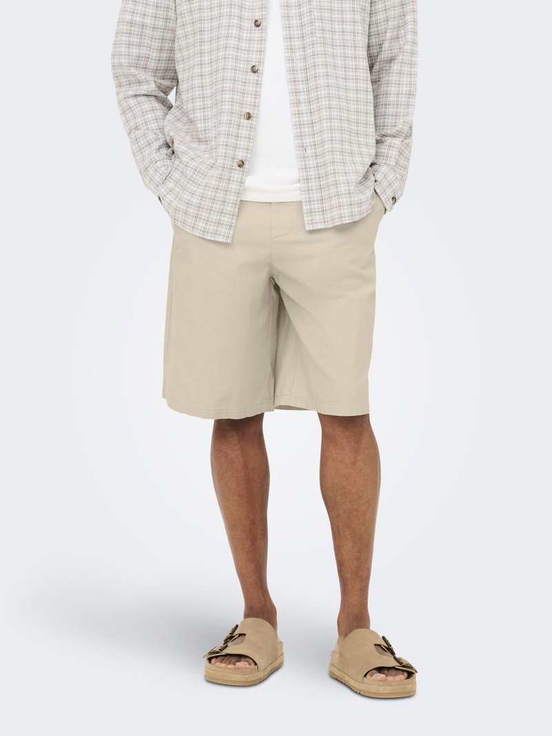 Homme portant un short beige mi-cuisses, des sandales ouvertes beiges avec des boucles, une chemise blanche et une chemise claire à carreaux boutonnée, les mains dans les poches.