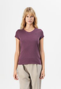 Blonde Frau, die ein enganliegendes, kurzärmeliges lila V-Ausschnitt-Shirt und lockere beige Hosen trägt, und vor einem weißen Hintergrund steht.