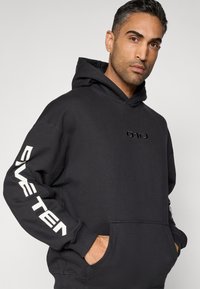 Sweatshirt preta com capuz, feita de tecido macio, com bolso frontal, capuz de cordão ajustável e logótipo branco e ousado "FIVE TEN" na manga e no peito.