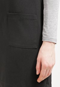 Handen vilar bredvid ett svart plagg med en fyrkantig ficka, iförd en ljusgrå långärmad tröja.