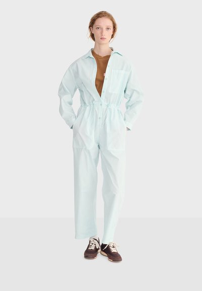 Jeune femme debout portant une combinaison bleu clair sur un t-shirt marron et des baskets foncées, sur un fond uni clair.