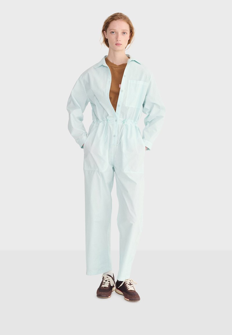 Jeune femme debout portant une combinaison bleu clair sur un t-shirt marron et des baskets foncées, sur un fond uni clair.