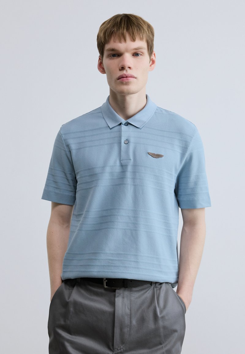 Ανοιχτό μπλε πόλο πουκάμισο με γιακά, με οριζόντια ριγέ υφή και ένα μικρό έμβλημα στο αριστερό στήθος. Συνδυάζεται με γκρι παντελόνι.