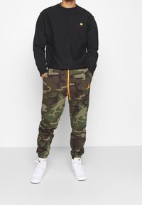 Svart sweatshirt med logotyp på bröstet, tillsammans med grön camo-mjukisbyxor med orange snörning och Jordan-logotyp. Vita sneakers fulländar stilen.