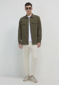 Uomo che indossa una camicia button-down verde scuro sopra una maglietta bianca, pantaloni beige chiaro, scarpe beige e occhiali da sole scuri, in piedi di fronte a uno sfondo neutro.