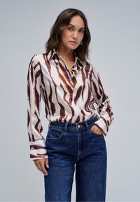 Frau trägt eine langärmelige braun-weiß gestreifte Bluse, in hoch taillierte dunkelblaue Jeans gesteckt, eine Hand in der Tasche, neutraler Hintergrund.