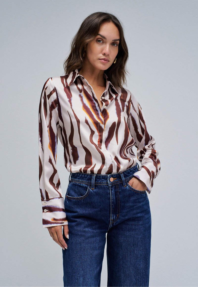 Frau trägt eine langärmelige braun-weiß gestreifte Bluse, in hoch taillierte dunkelblaue Jeans gesteckt, eine Hand in der Tasche, neutraler Hintergrund.