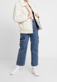 Kræmfarvet, fuzzy jakke med krave, parret med højtaljede blå denim cargo bukser. Hvide sneakers med en tekstureret sål.